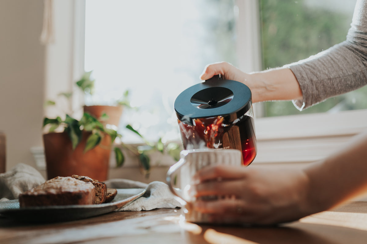 Woman pouring coffee from a Moccamaster glass carafe into a ceramic mug.