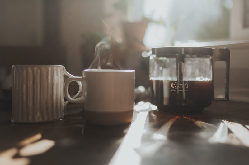 two white mugs sit on a sunbeam-lit surface, steaming. to the right, a kbg carafe, half full and steaming as well.