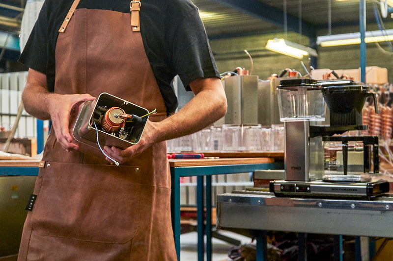 A leather aproned worker, face off-screen, holds the main tower of a moccamaster with boiling element exposed. A complete moccamaster, as well as a factory-worth of parts is framed in the background