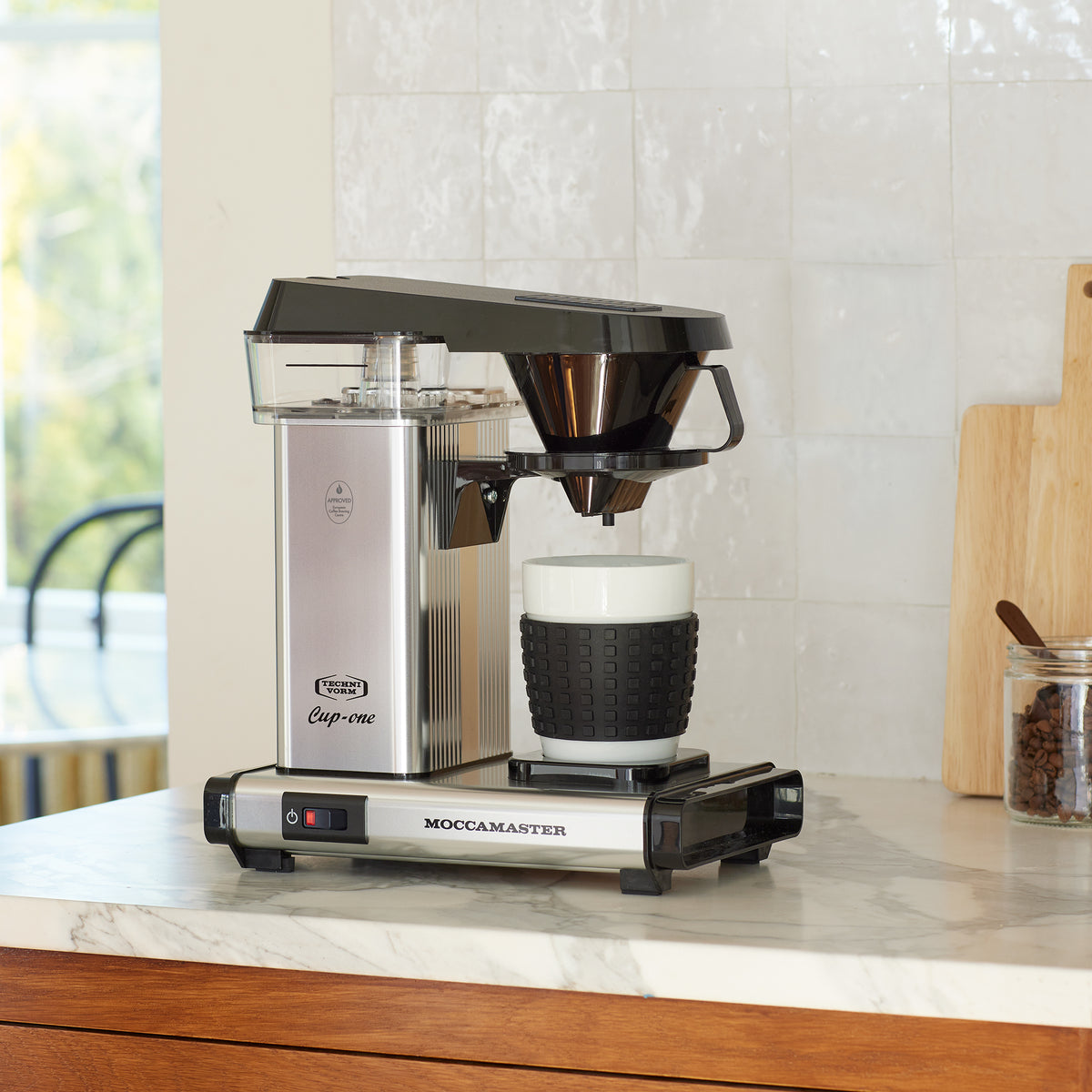 Silver Cup-one single-cup moccamaster on a marble counter with white tile backsplash, with a cuttingboard and a jar of coffeebeans with a coffee scoop in the background.
