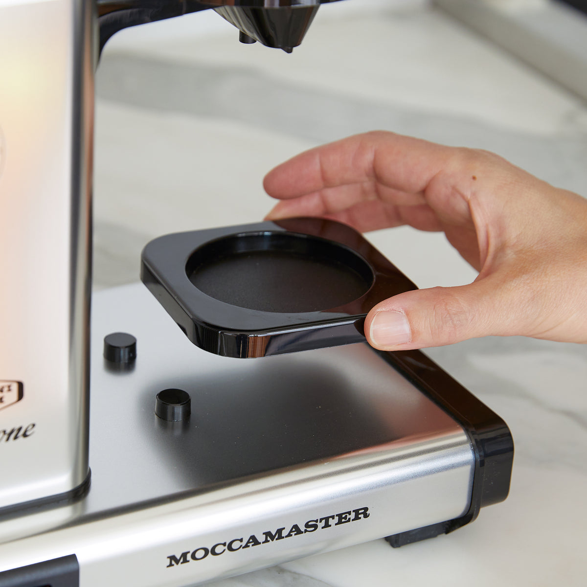 close-up of the cup-one, a hand is holding the removable cup holder to show the plastic posts underneath, where a variety of cup sizes can fit. Moccamaster badging is on the body of the brewer.