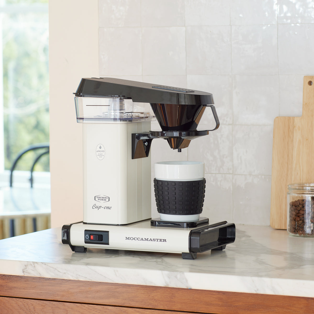 off-white Cup-one single-cup moccamaster on a marble counter with white tile backsplash, with a cuttingboard and a jar of coffeebeans with a coffee scoop in the background.
