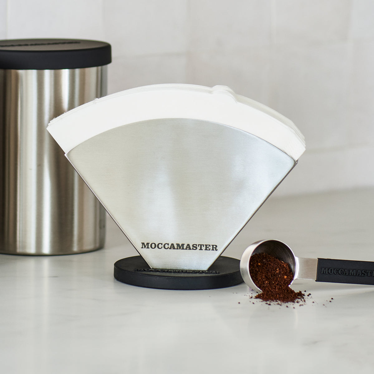 Close up shot of Moccamaster stainless steel accessories, including a coffee bean canister, paper filter holder, and coffee ground scoop.
