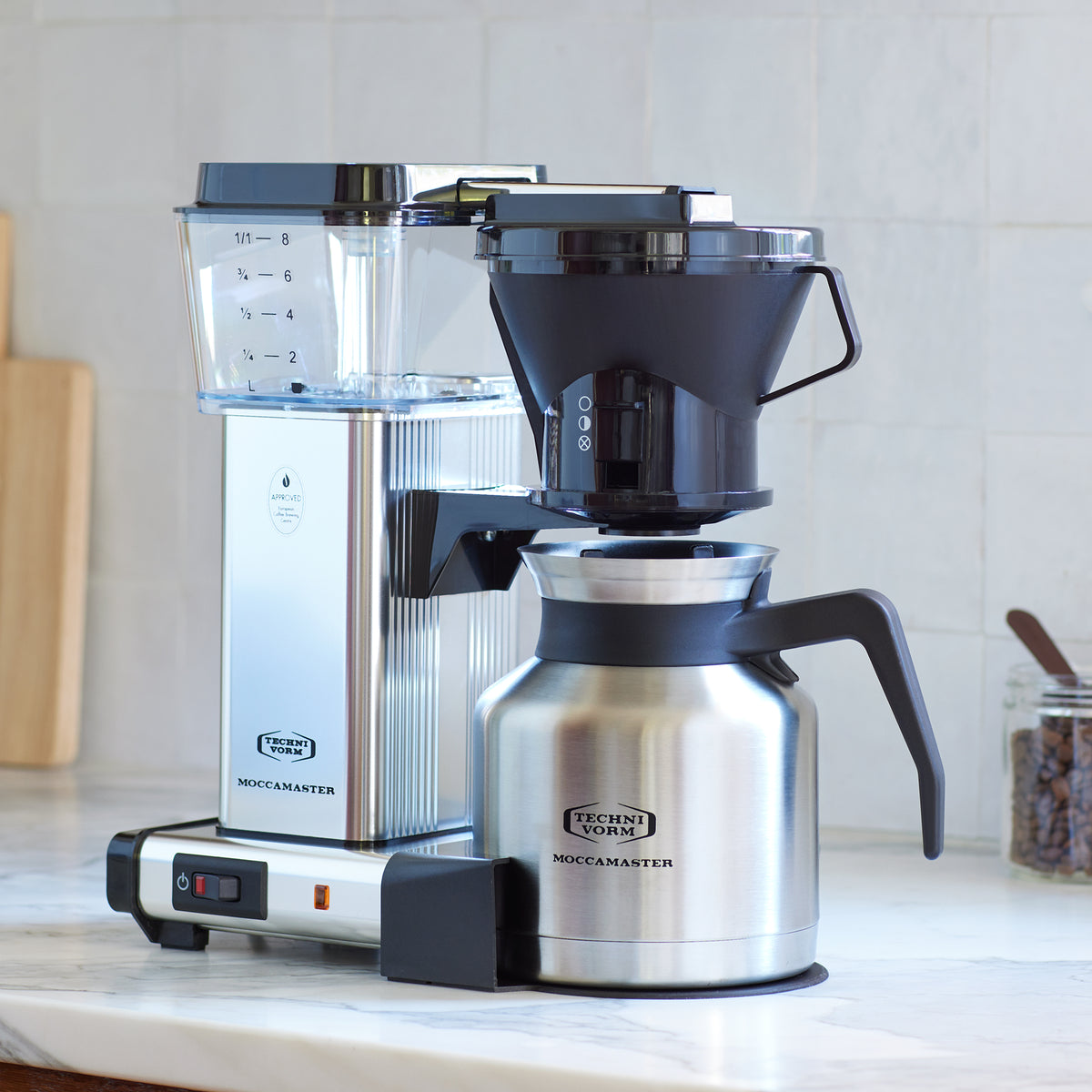 Moccamaster KBTS in Polished Silver, on a marble counter with white tile backsplash  A jar of coffee beans with a scoop on the right, a wooden cutting board on the left, in the background.