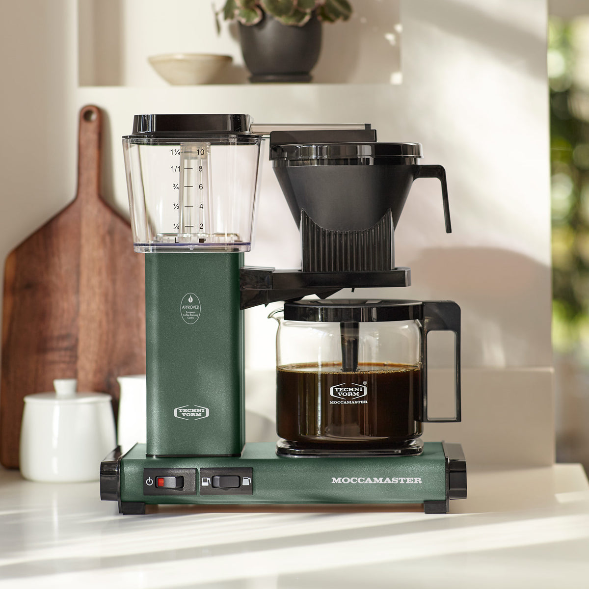 Green Moccamaster coffee maker on a countertop, beside a wooden board, with a plant in the background.