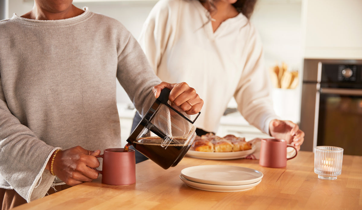 Two people standing in a kitchen - one pours coffee while another one is about to pick up a tray of sticky buns.