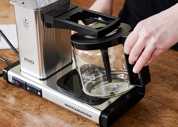 Person placing a glass carafe into a Moccamaster KBGV Select coffee brewer.