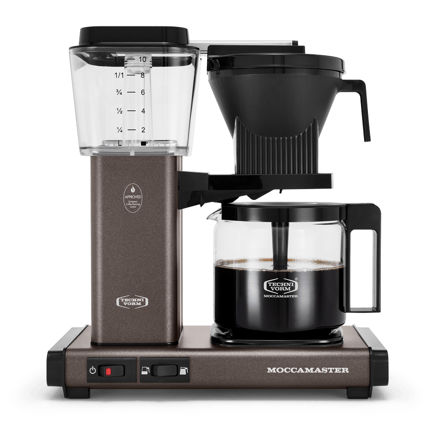 Front shot showing Moccamaster KBGV Select in Stone Grey, with rectangular tower and base, clear acrylic water reservoir with fill level marks, power and volume selector switch, glass carafe with black handle, and black automatic brew basket.