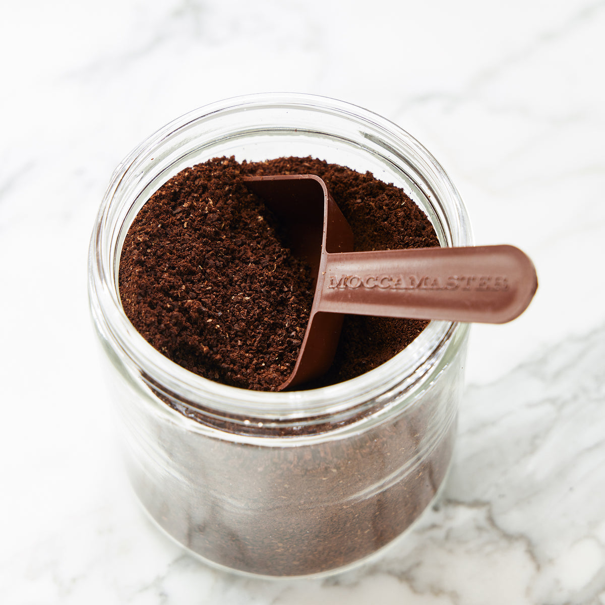 A brown 2-tablespoon Moccamaster coffee scoop inside a glass jar full of coffee grounds.