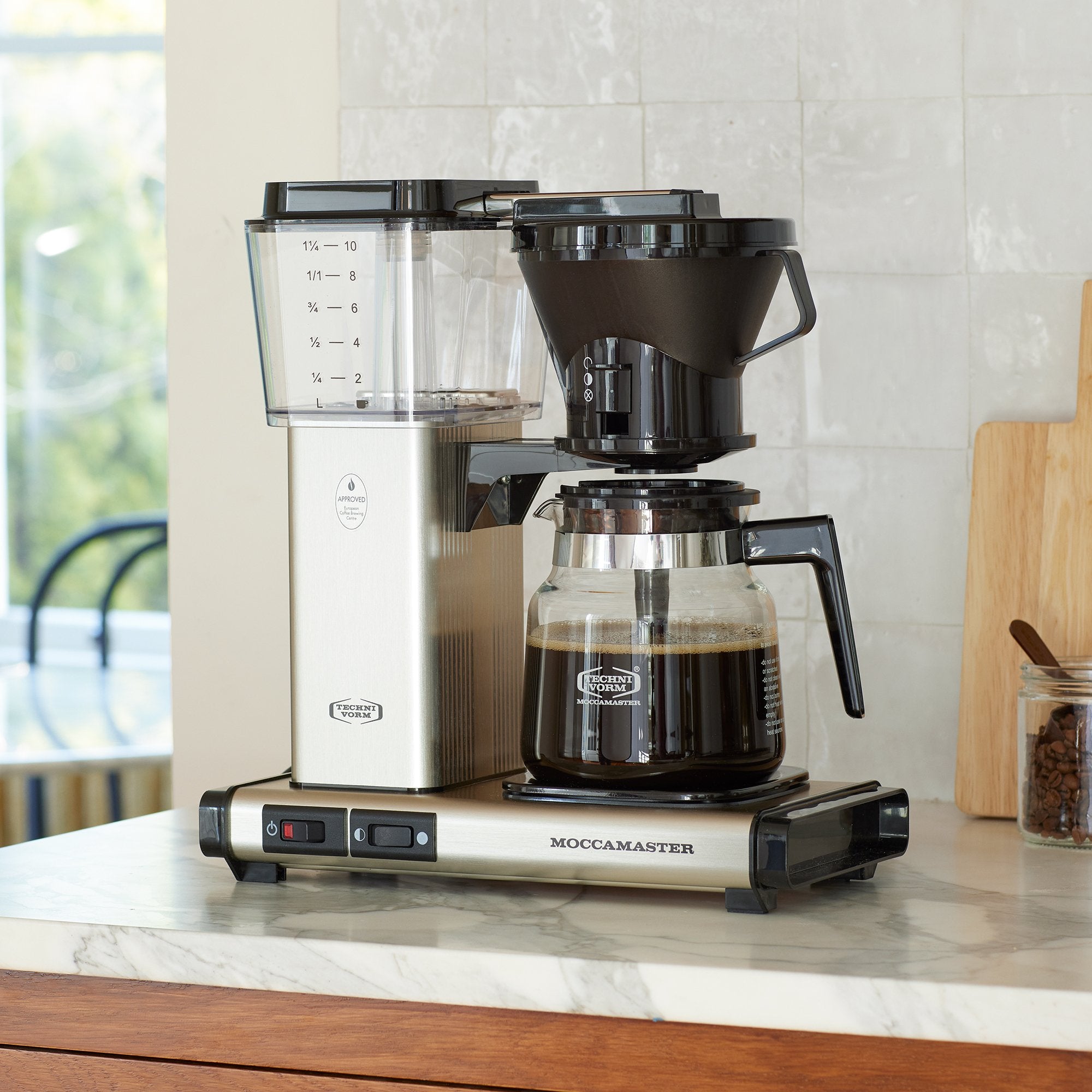 Moccamaster KB in Brushed Silver -Front shot showing rectangular tower and base, clear acrylic water reservoir with fill level marks, power and hotplate level switches, glass carafe with plastic handle, and black brew basket with drip speed selector.