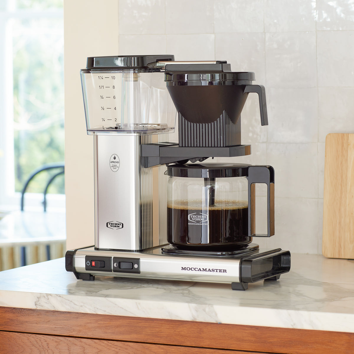 Moccamaster KBG in Polished Silver, on a marble counter with white tile backsplash, with a cutting board and a jar of coffee beans with a coffee scoop in the background.