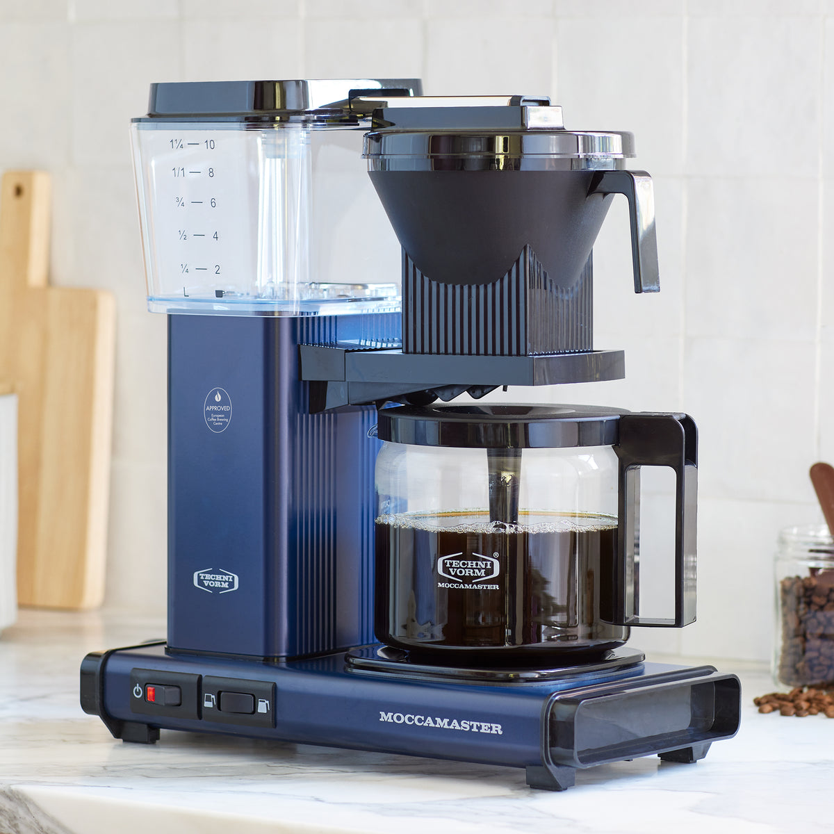 Moccamaster KBGV Select in Midnight Blue, on a marble counter with white tile backsplash. A jar of coffee beans with a scoop on the right, a wooden cutting board on the left, in the background.