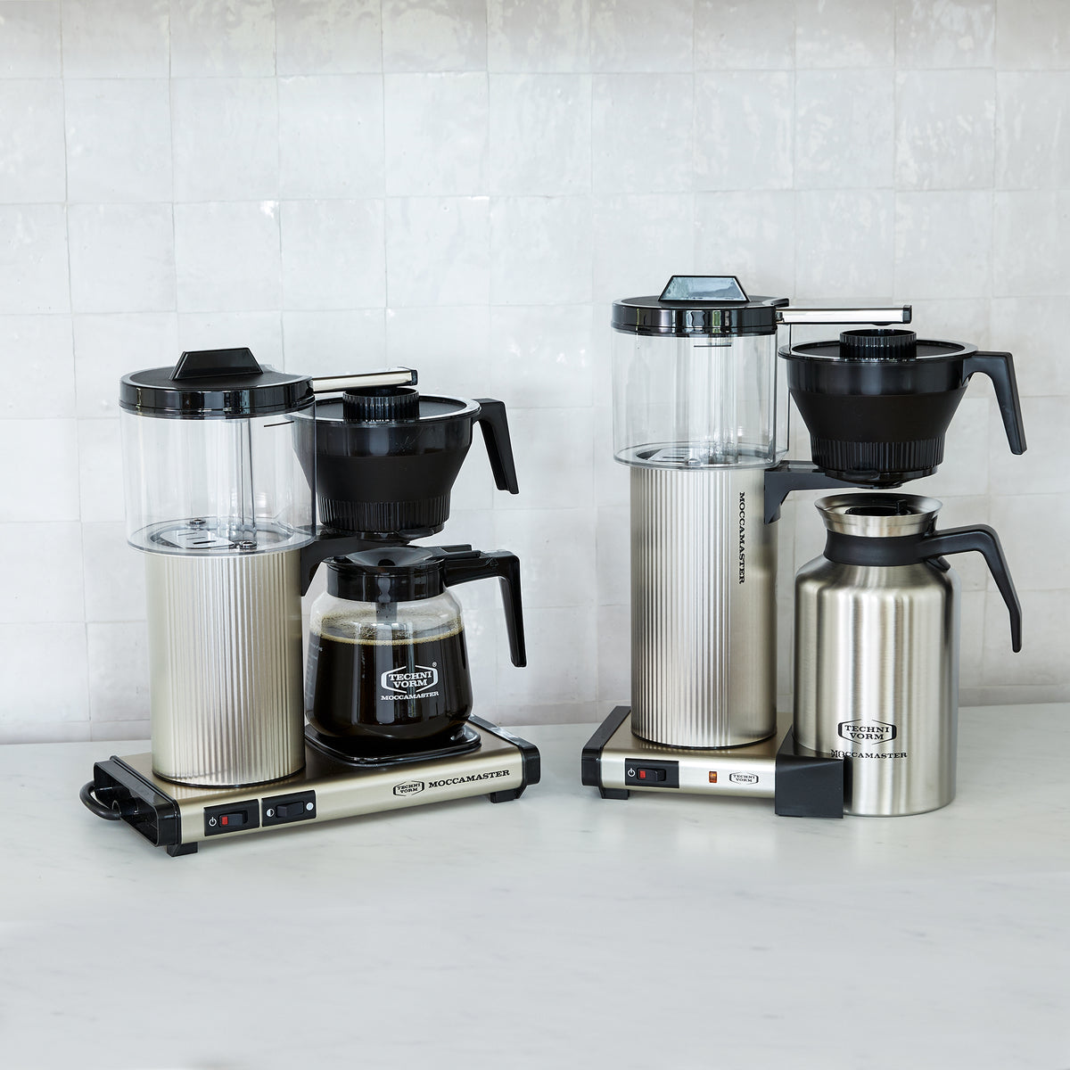 Collection of Moccamaster coffee machines with various designs, including clear water reservoirs, silver bodies, glass carafes, and stainless steel carafes, displayed against a white tiled backdrop.