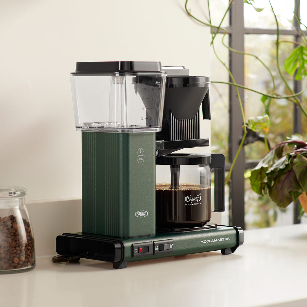 A green Moccamaster coffee machine on a counter, with a glass carafe, water tank, and adjacent coffee beans.