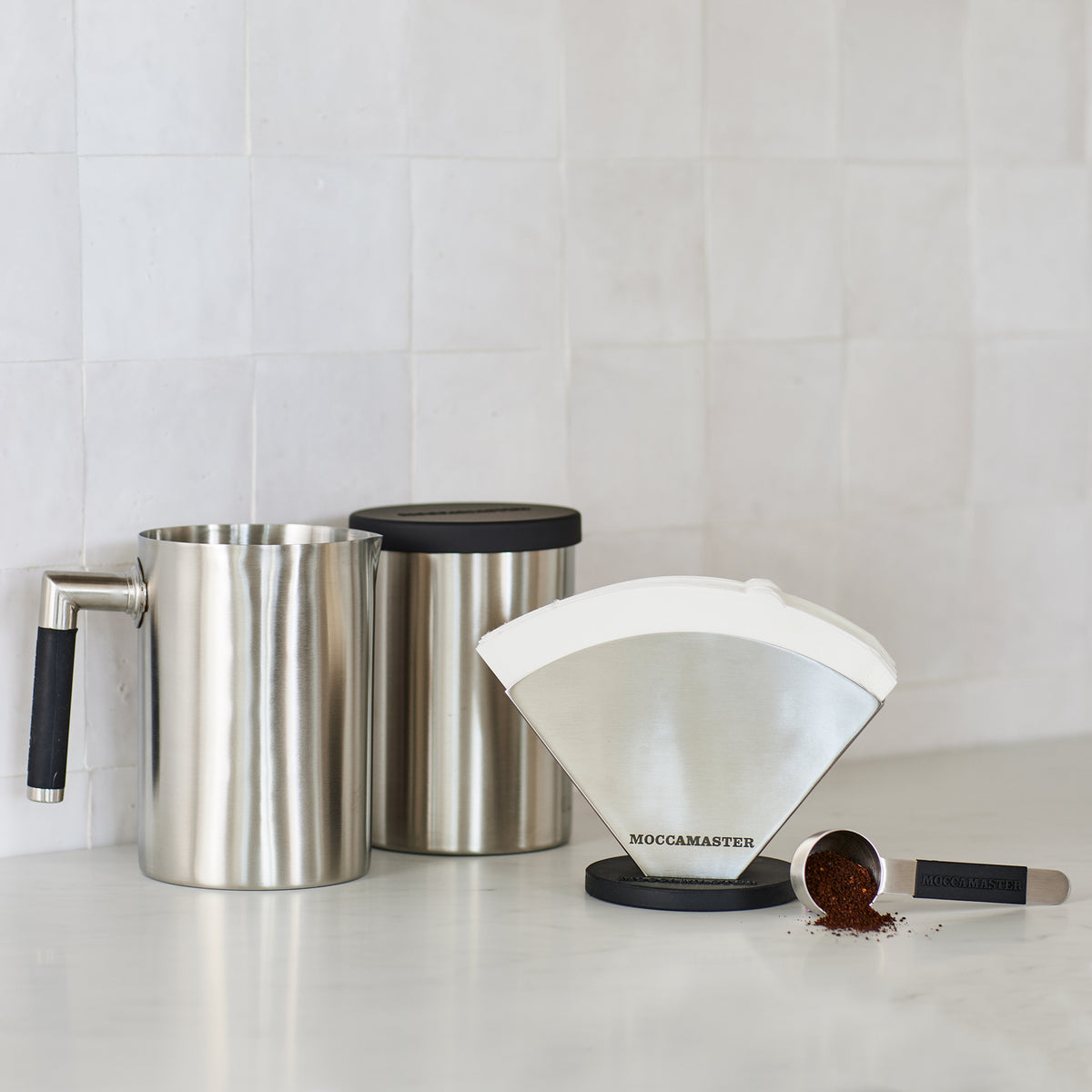 A collection of stainless steel Moccamaster accessories, including a filter holder, coffee scoop, coffee canister, and water jug.