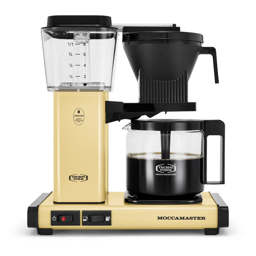 Front shot showing Moccamaster KBGV Select in Butter Yellow, with rectangular tower and base, clear acrylic water reservoir with fill level marks, power and volume selector switch, glass carafe with black handle, and black automatic brew basket.