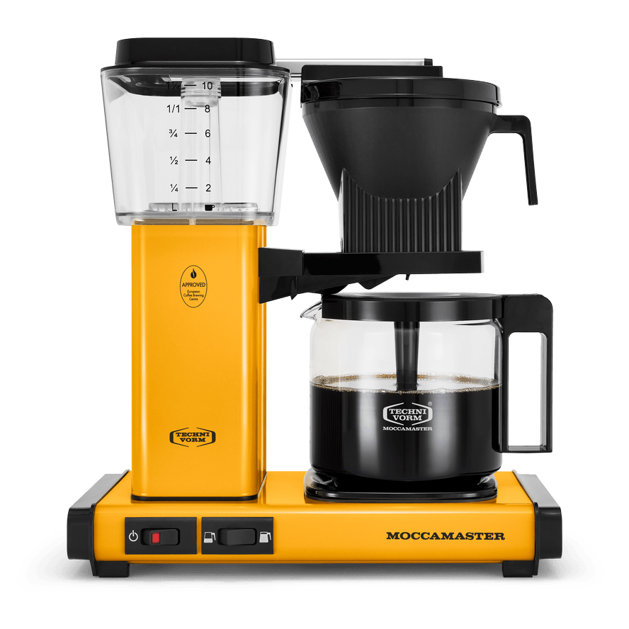 Front shot showing Moccamaster KBGV Select in Yellow Pepper, with rectangular tower and base, clear acrylic water reservoir with fill level marks, power and volume selector switch, glass carafe with black handle, and black automatic brew basket.