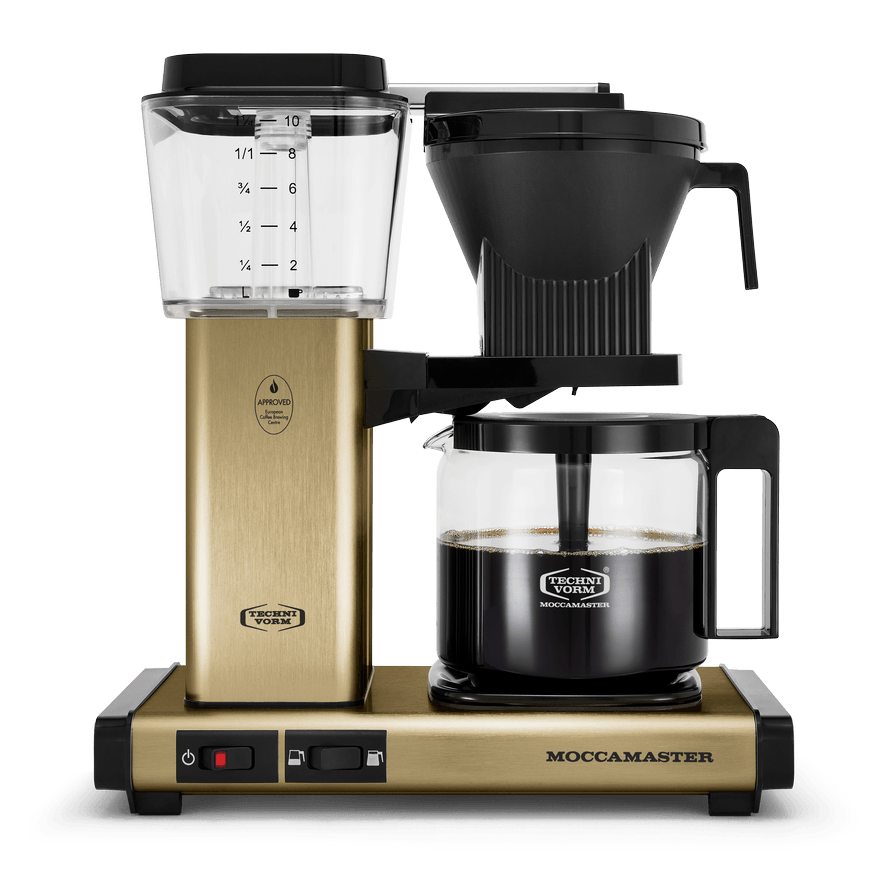 Front shot showing Moccamaster KBGV Select in Brass, with rectangular tower and base, clear acrylic water reservoir with fill level marks, power and volume selector switch, glass carafe with black handle, and black automatic brew basket.